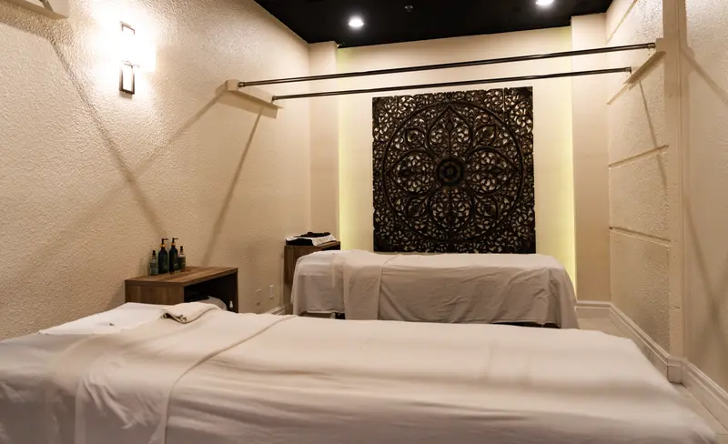 A couples room: two massage tables in white linen beneath a carved teak Thai panel