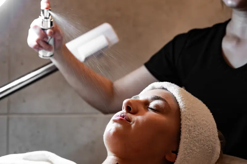 A Massage 1 esthetician uses a facial steamer over a client lying back with a towel wrapped around her hair